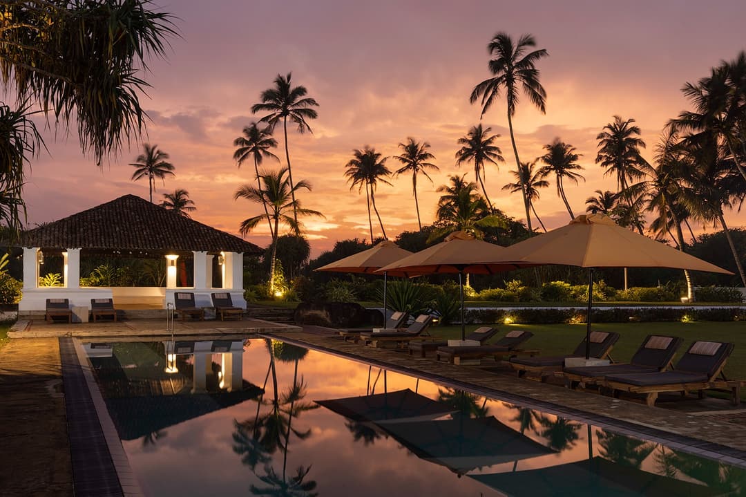 Sunset over Heritage House Bentota pool with coconut palms and illuminated colonial building